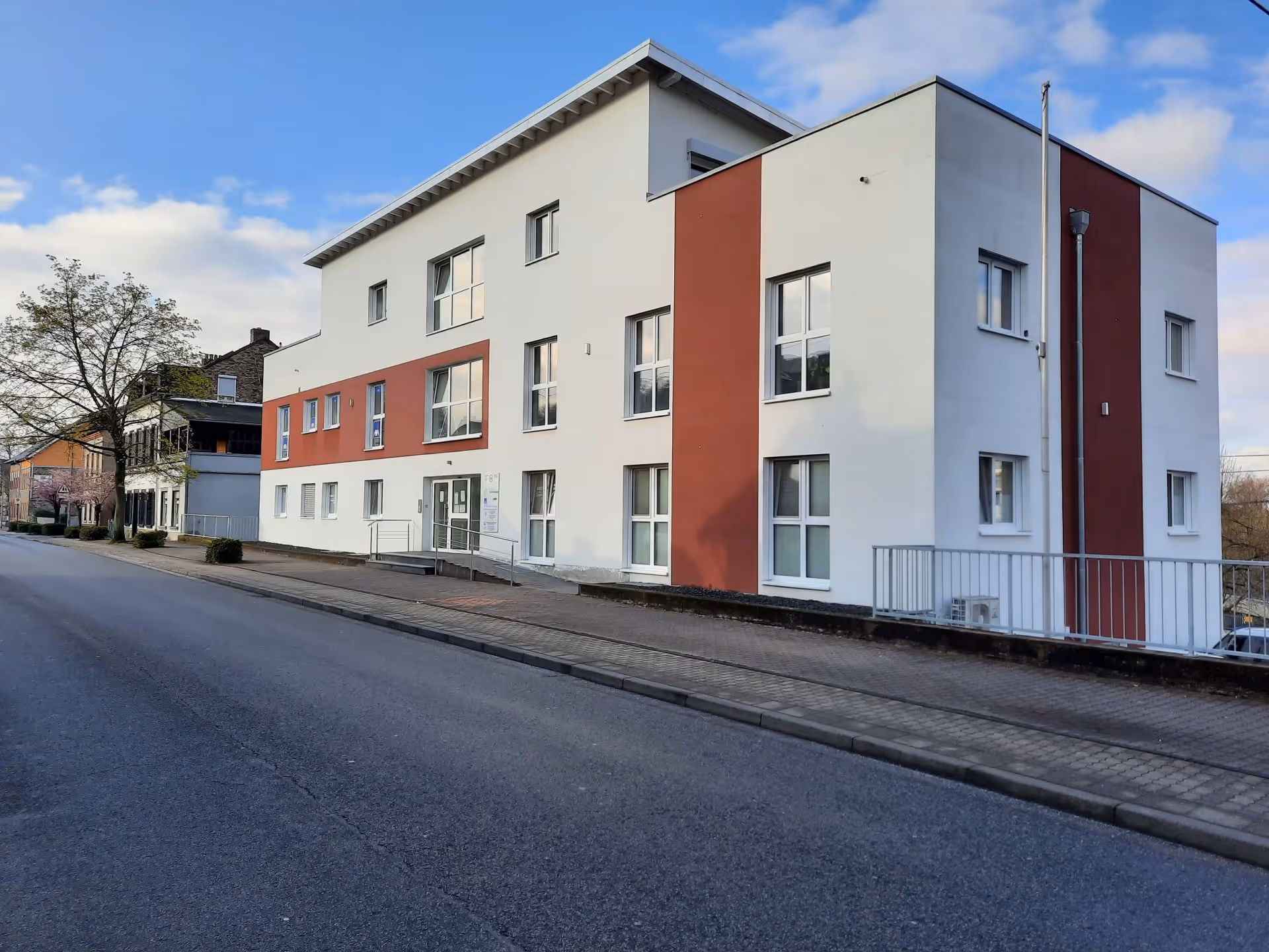 Three-story white building with red vertical accents along a quiet street on a partly cloudy day.