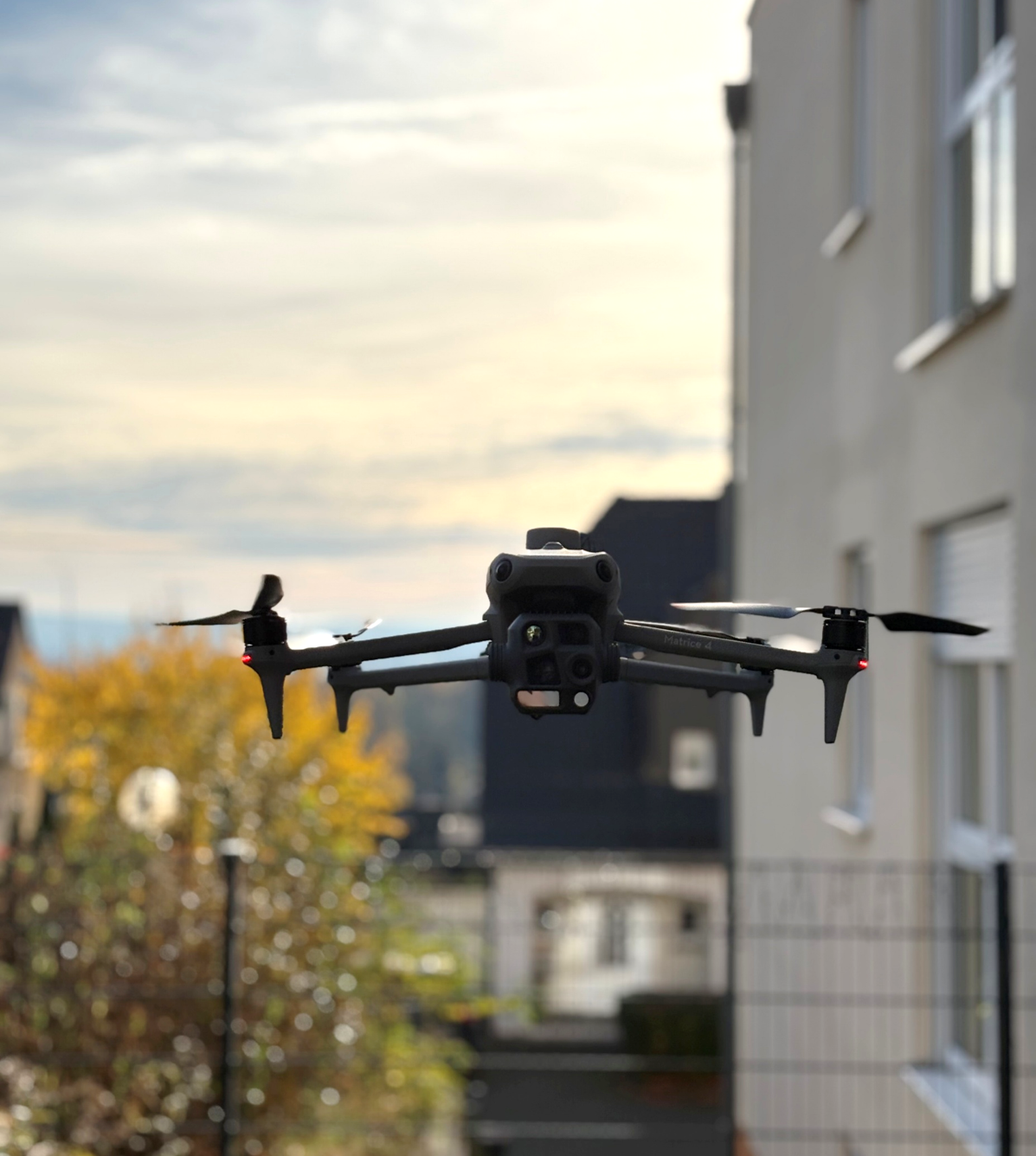 Black DJI Matrice 4 drone hovering outdoors with blurred houses and autumn foliage in the background.