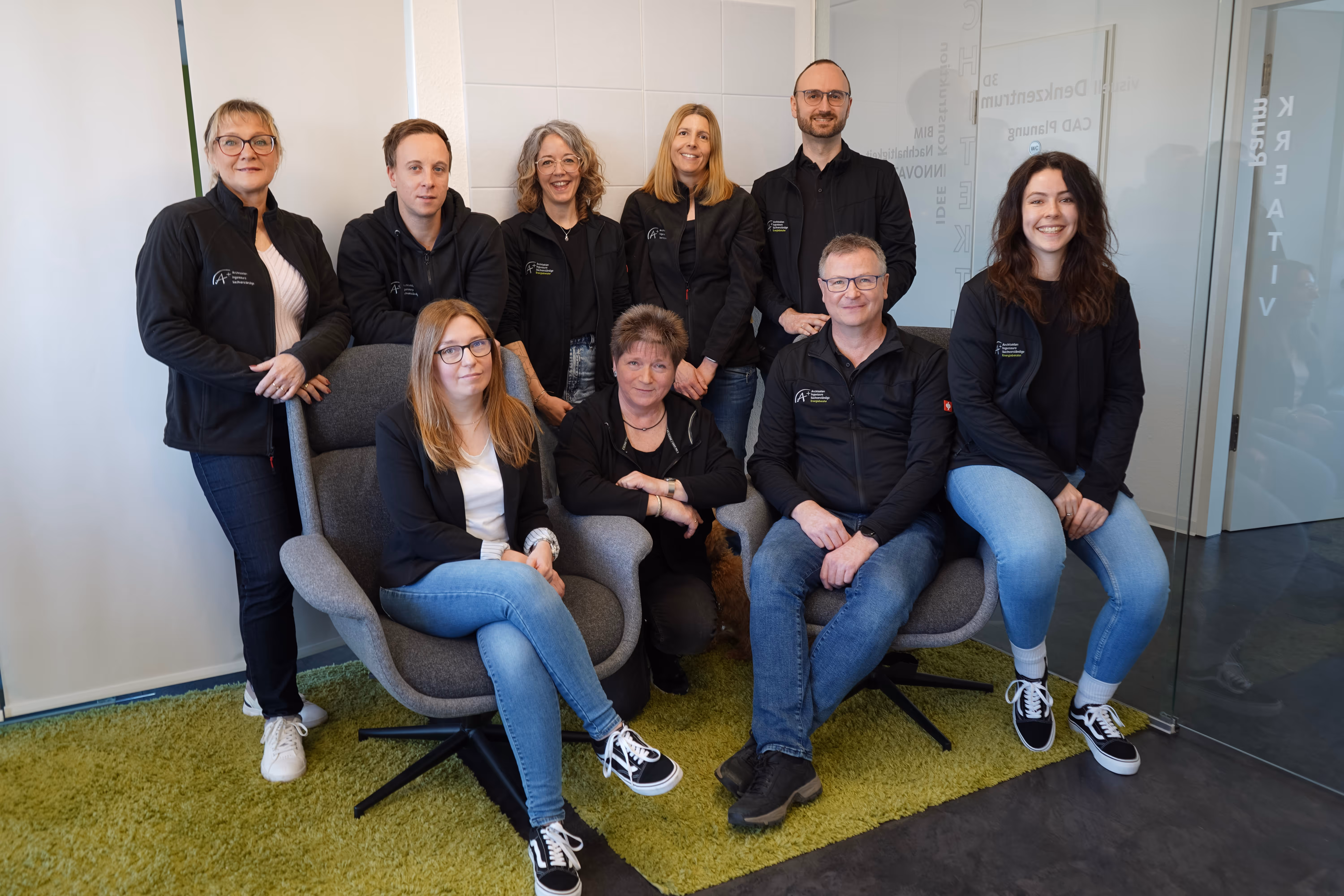 Group of nine smiling adults in casual black jackets posing in an office setting with two gray armchairs on green carpet.