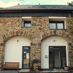 Stone building facade with two arched doorways, one with a wooden bench outside and a potted plant by the other entrance.