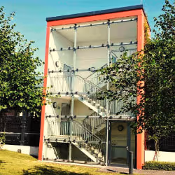 Three-story outdoor fire escape staircase enclosed in glass with a red frame, surrounded by trees and grass.