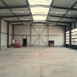 Empty industrial warehouse interior with concrete floor and metal frame walls, featuring an overhead skylight and large doors.