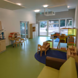 Bright, spacious preschool classroom with green floor, wooden furniture, toys, and large windows letting in natural light.