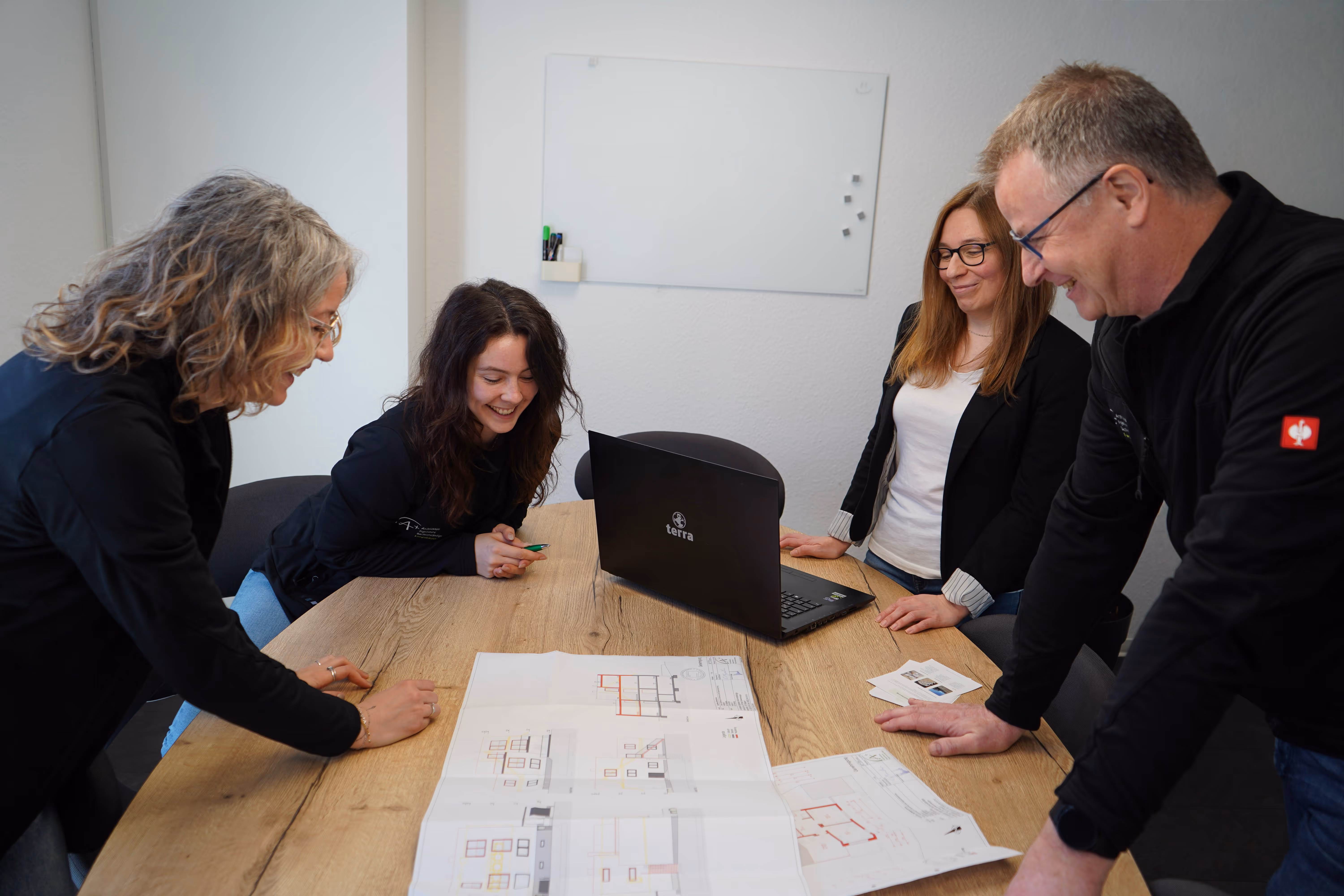 Four people smiling and discussing architectural plans on a wooden table with a laptop in a meeting room.