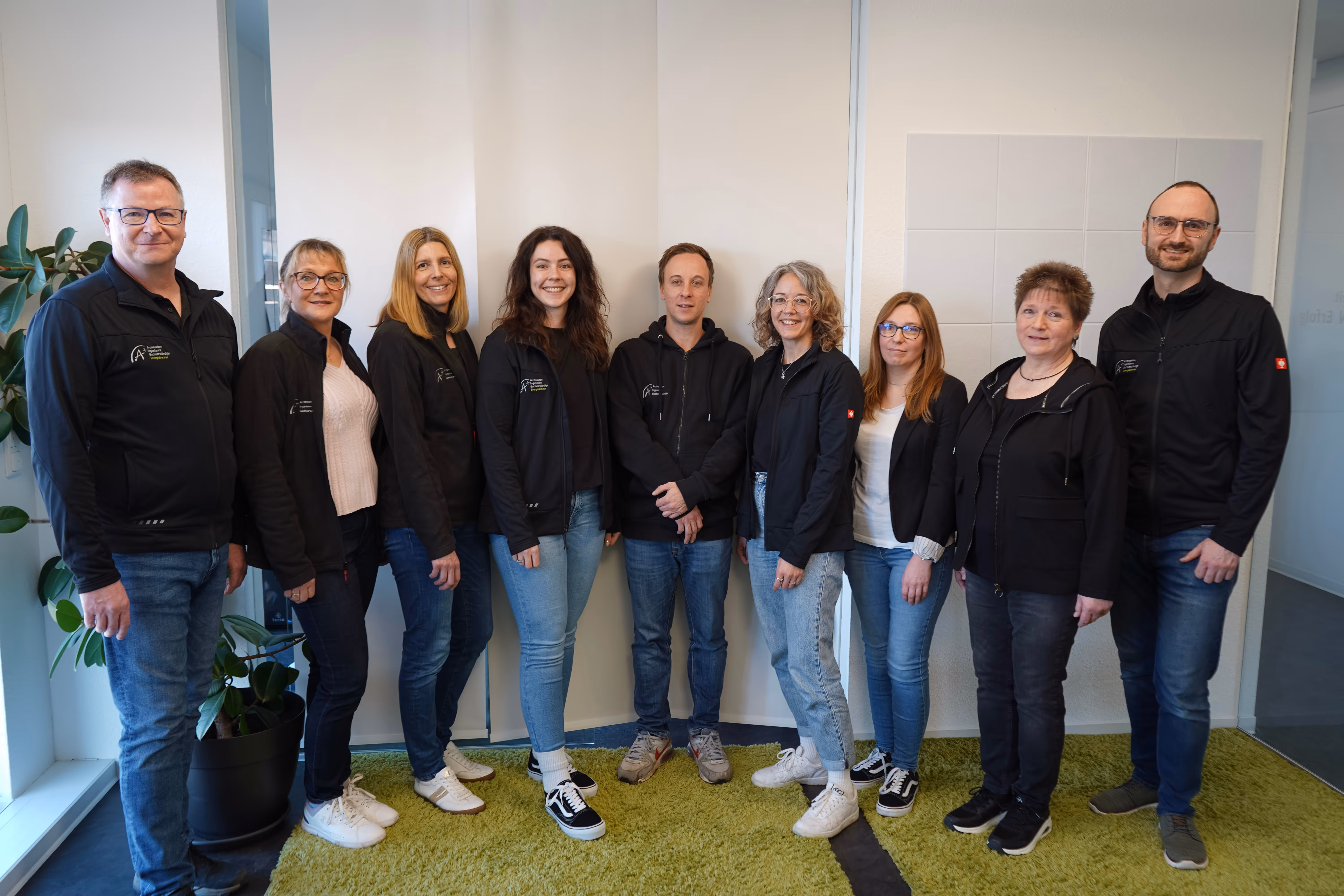 Group of nine adults standing in a row indoors, wearing casual clothes and black jackets, smiling at the camera.